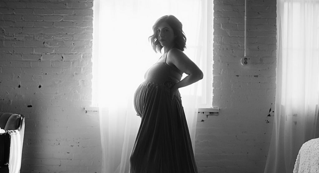 Black and White image of a pregnant woman posing in front of a curtained window wearing a maxi dress and holding her baby bump