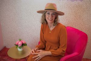 Kristin Revere of Gold Coast Doulas sits on a pink chair in an orange dress and hat