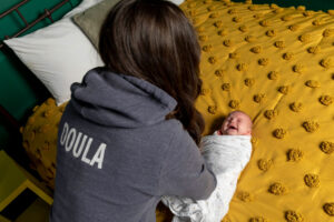 Kay from Gold Coast Doulas wearing a Doula hoodie, swaddling a baby on a yellow bed