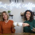 Kristin & Alyssa from Gold Coast Doulas cheers two coffee mugs in front of a neon sign that says "but first, coffee"