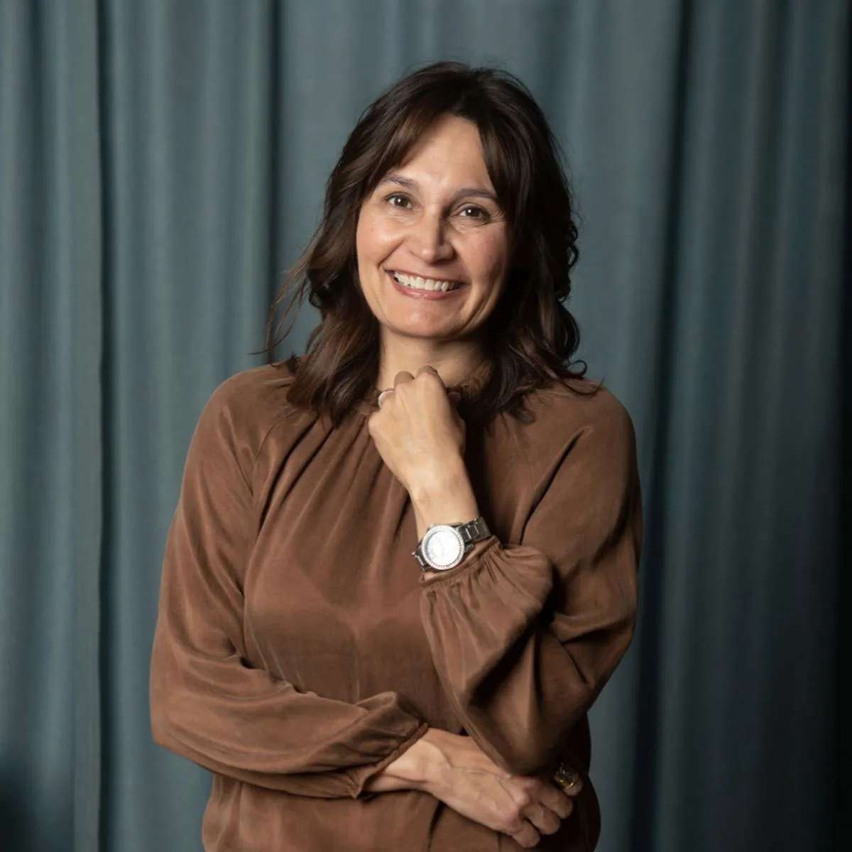 Lisa Newhouse smiling for a photo wearing a brown shirt in front of a grey curtain
